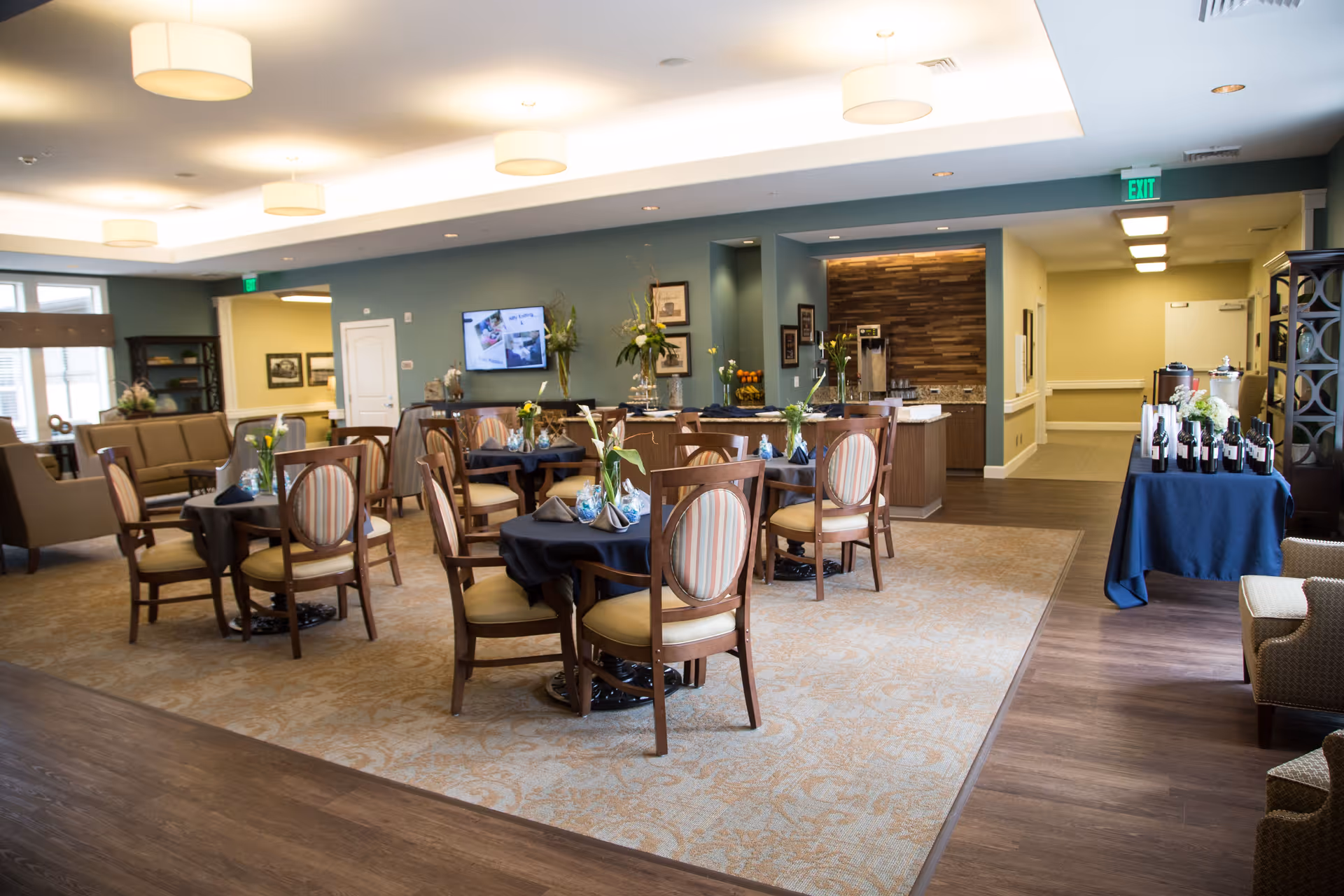 A spacious dining area in a senior living facility with several round tables covered with dark tablecloths, each set with napkins and small flower arrangements. The room has comfortable wooden chairs with cushioned seats and backs, a carpeted floor area surrounded by wood flooring, and soft overhead lighting. In the background, there is a small kitchenette or serving area with a coffee machine and a hallway leading further into the facility. A television is mounted on the wall, and there are decorative plants and framed pictures on the walls.