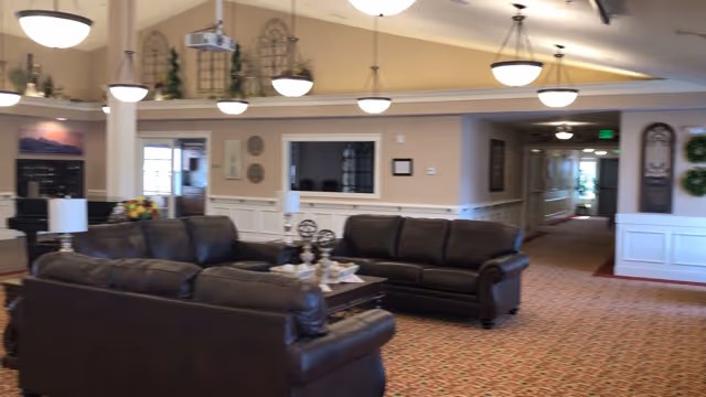 A spacious living room area in an assisted living facility featuring dark leather sofas arranged around a coffee table with decorative items. The room has beige walls, patterned carpet, multiple ceiling lights, and decorative wall accents. A hallway extends from the living area towards the back.
