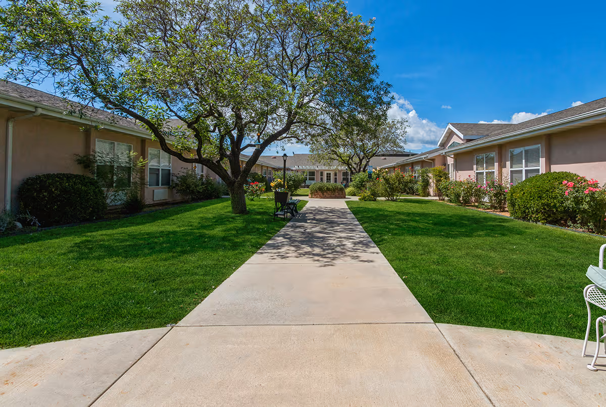 A sunlit landscaped courtyard with a central concrete walkway, trees, benches, and single-story senior living buildings on either side.