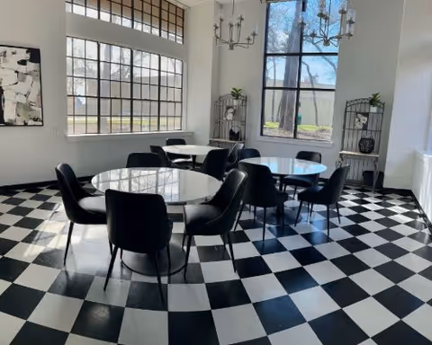 Bright communal dining area with round glass tables, black chairs, chandeliers, and a black-and-white checkered floor.