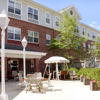 Outdoor patio area at a senior living facility with several tables and chairs, a swing with a canopy, potted plants, and a brick building with multiple windows in the background under a clear blue sky.