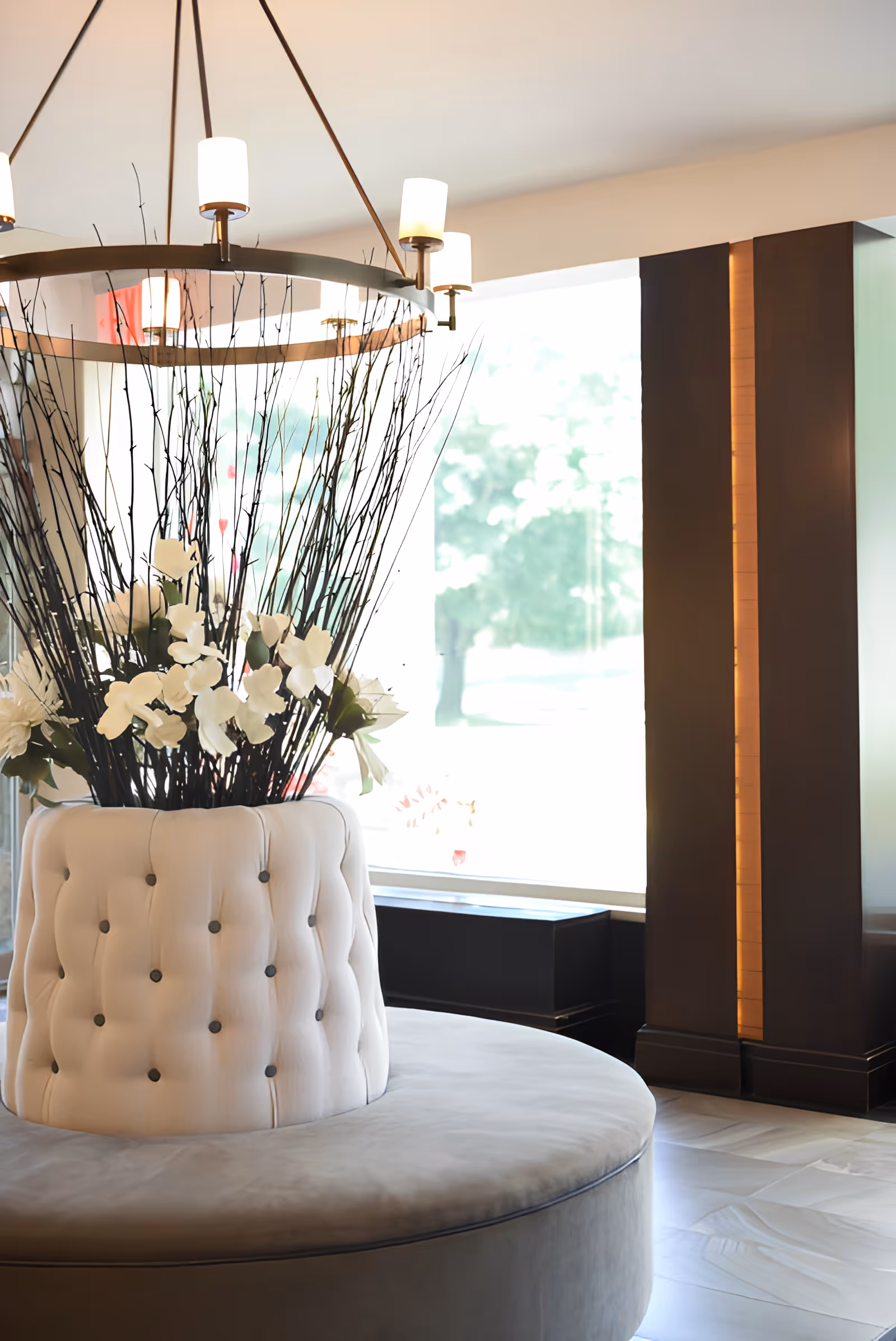 A modern circular seating area with tufted white upholstery surrounding a large floral arrangement of white flowers and tall branches, situated in a bright room with large windows and a contemporary chandelier overhead.
