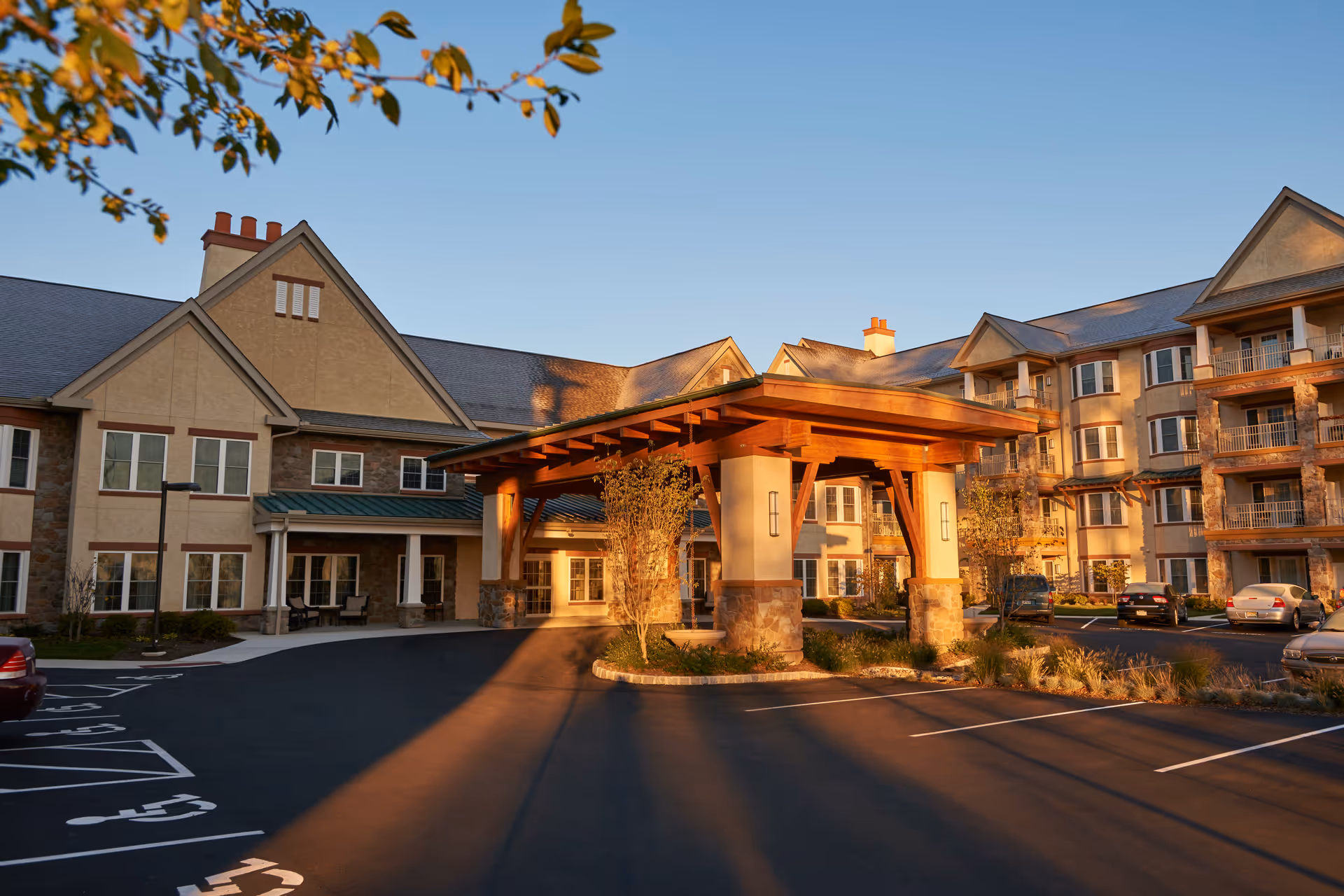 Exterior view of Tel Hai Retirement Community building during sunset, showing a large covered entrance with wooden beams and stone pillars, multiple windows, balconies, and a parking lot with several cars.