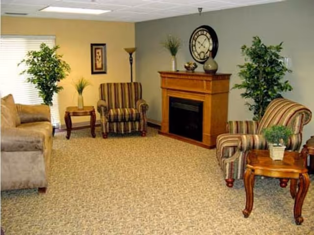 A cozy living room with a beige couch on the left, two striped armchairs, a wooden fireplace with decorative items and a clock above it, two wooden side tables with potted plants, a floor lamp, framed artwork on the wall, and two large green plants near the walls.