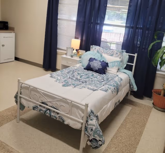 White metal bed with blue bedding and nightstand in a bright assisted living bedroom.