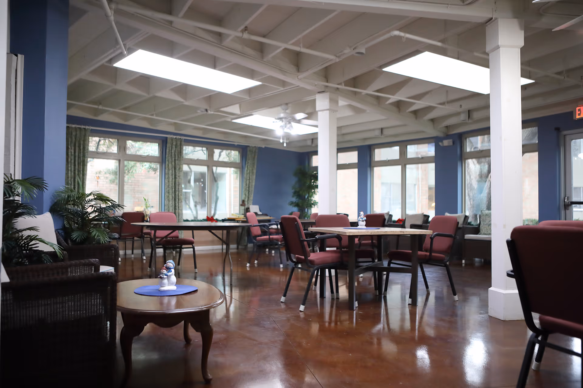 A spacious common area with multiple tables and chairs arranged for seating. The room has large windows with green curtains, blue walls, and a polished brown floor. There are some plants and small decorative items on the tables. The ceiling has exposed beams and skylights, providing natural light.