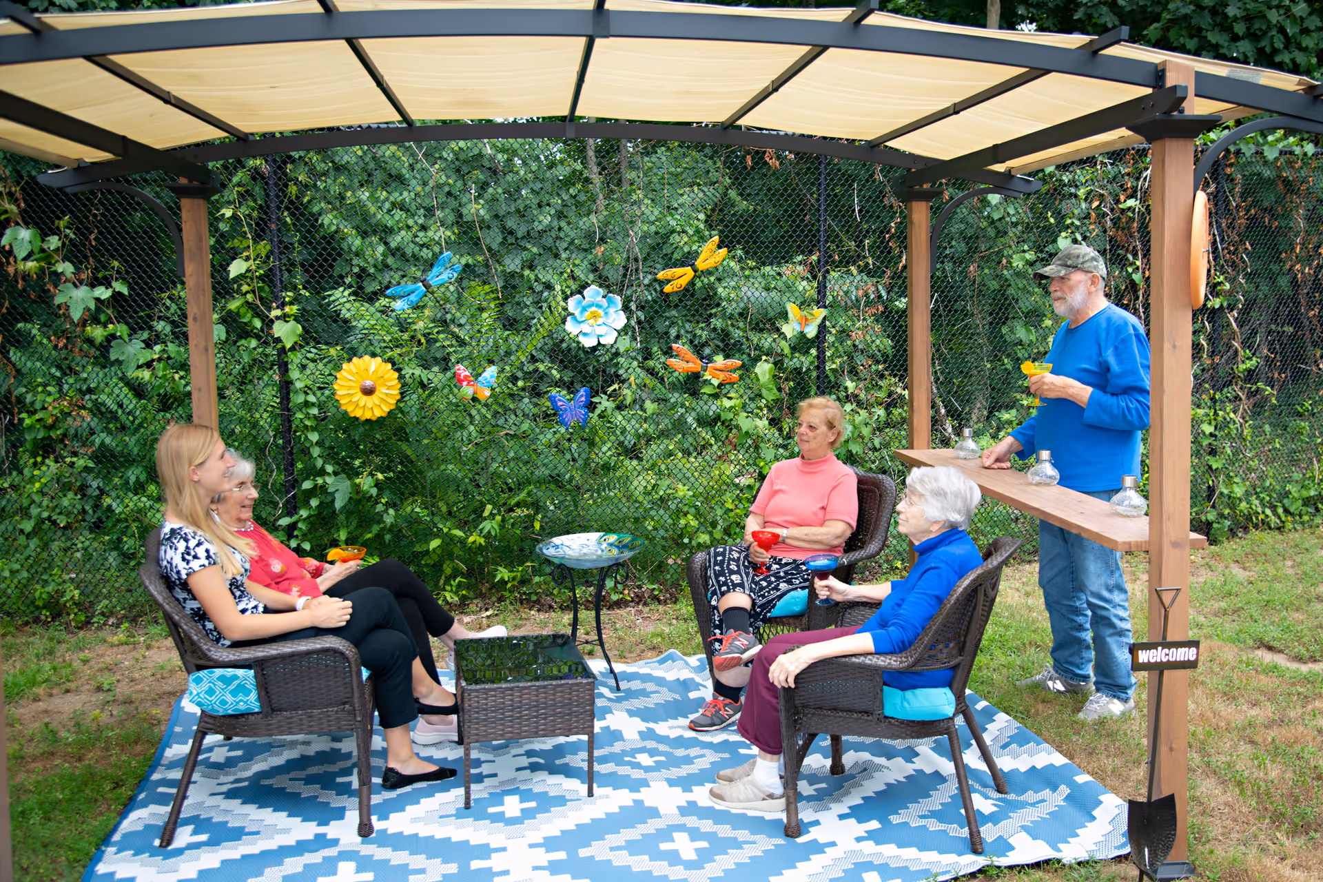 Five seniors sitting and standing under a shaded outdoor pergola with a blue and white patterned rug, wicker chairs, and a small table. Colorful butterfly and flower decorations hang on a chain-link fence covered with greenery in the background. One man is standing holding a drink, while four women are seated, also holding drinks, enjoying a social gathering in a garden setting.
