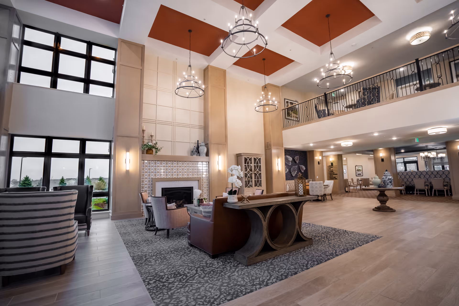 Spacious and elegant senior living facility common area with high ceilings, large windows, modern chandeliers, a fireplace, comfortable seating including armchairs and sofas, decorative vases, and a mezzanine level overlooking the space.