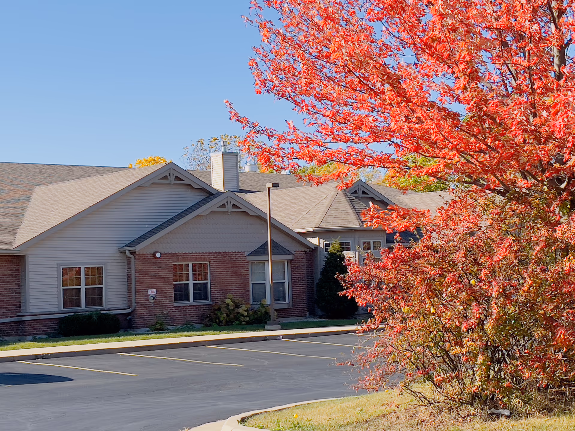 Exterior view of a single-story building with brick and siding walls, a shingled roof, and several windows. In the foreground, there is a parking lot with marked spaces and a vibrant tree with bright red autumn leaves on the right side. The sky is clear and blue.