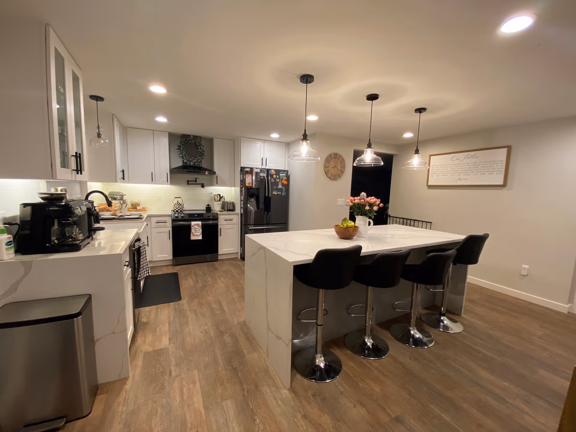 Modern kitchen with white cabinets, black appliances including a refrigerator and stove, a large white island with four black bar stools, three pendant lights hanging above the island, wooden flooring, and a wall clock and framed text on the wall.