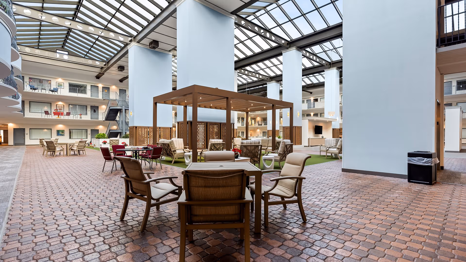 Spacious indoor common area with a high glass ceiling, multiple seating arrangements including tables and chairs, a wooden pergola structure with cushioned chairs underneath, and surrounding balconies with doors and windows overlooking the space.