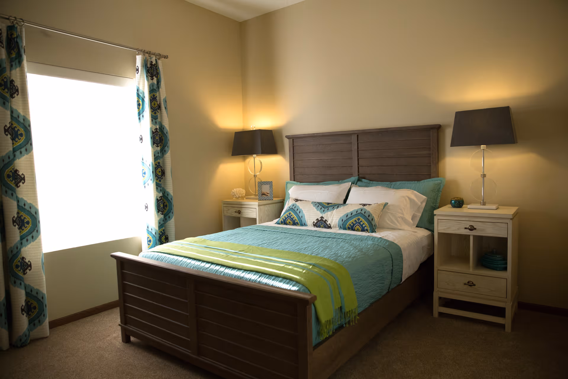 A cozy bedroom with a wooden bed frame and headboard, neatly made with teal and white bedding and decorative pillows. There are two matching nightstands on either side of the bed, each with a lamp and small decorative items. A window with patterned curtains allows natural light to brighten the room.