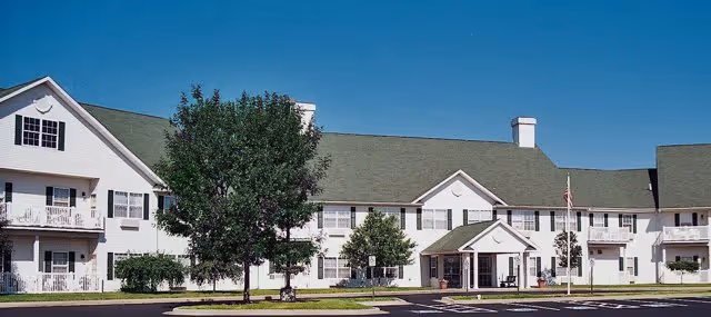 Exterior view of a large, two-story senior living facility with white siding and a green roof under a clear blue sky. The building has multiple windows, balconies, and a covered entrance with an American flag on a flagpole nearby. There are trees and shrubs in front of the building and a paved parking area in the foreground.