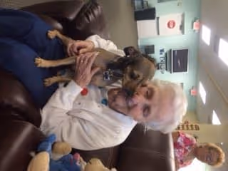 An elderly woman sitting on a brown leather couch holding a small brown dog. The woman is smiling and wearing a white sweater. In the background, there is another person and a room with light blue walls and ceiling lights.