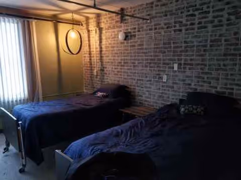A dimly lit bedroom with two single beds covered in dark blue bedding. The room has an exposed brick wall, a window with vertical blinds, and a circular mirror hanging on the wall near the window. There is a small wooden bench between the beds.