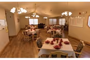 Communal dining room with multiple tables set with red place settings, wooden chairs, chandeliers, and windows.