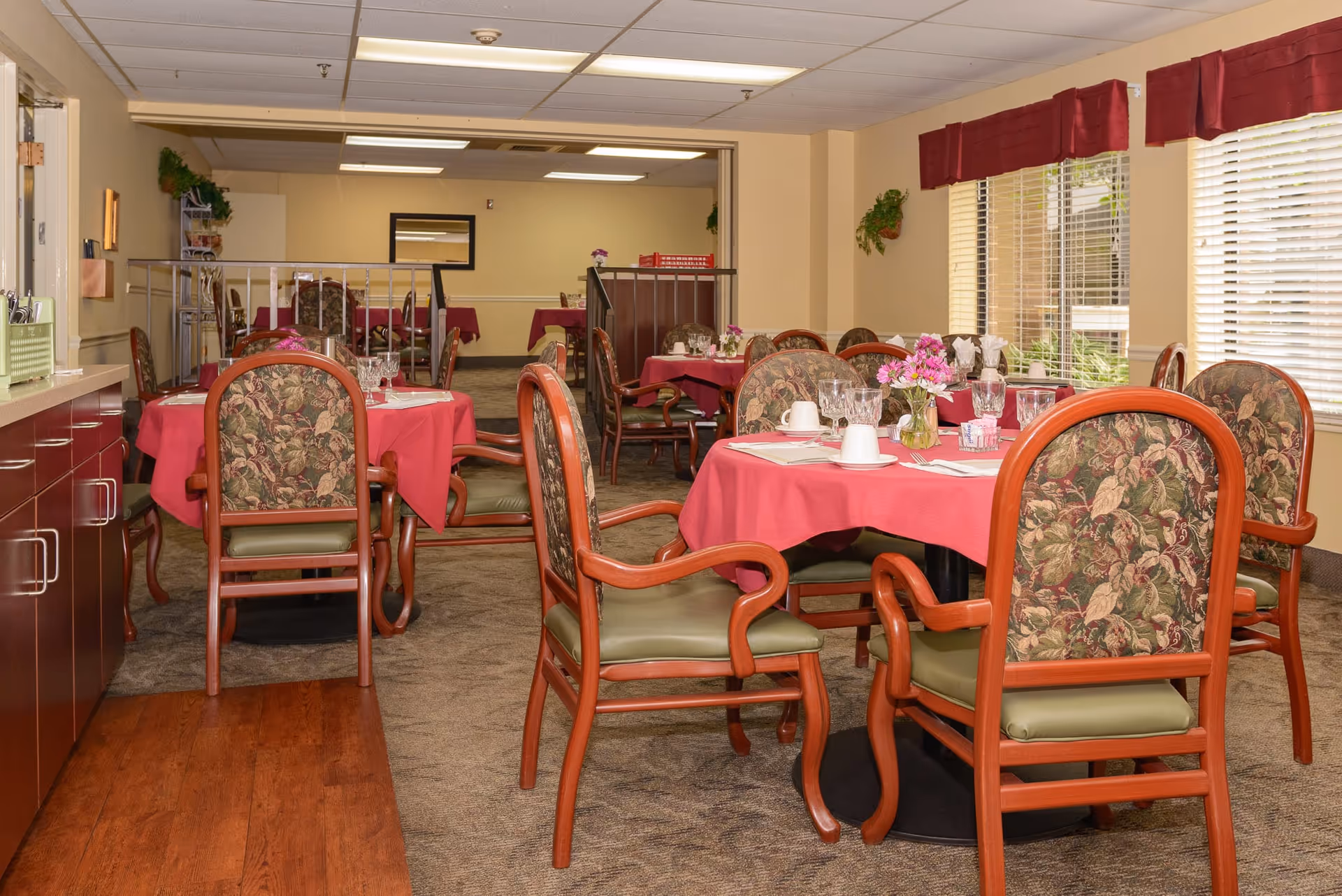 Dining room in a senior living facility with round tables covered in red tablecloths, floral centerpieces, and chairs with floral upholstery and wooden frames. Large windows with blinds allow natural light into the room.