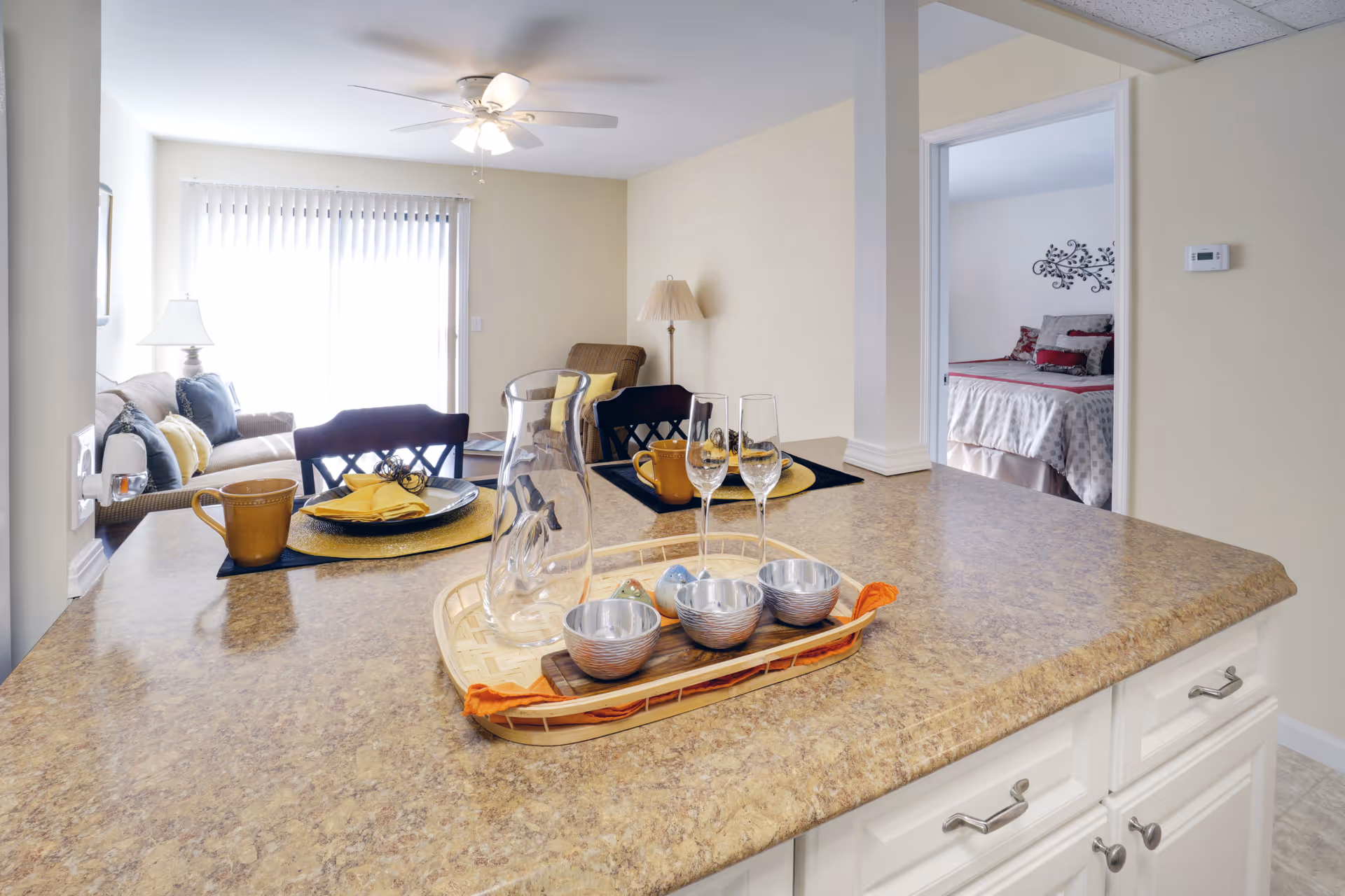 Open-plan interior with a kitchen island set with dishes and glassware overlooking a living room and a bedroom.