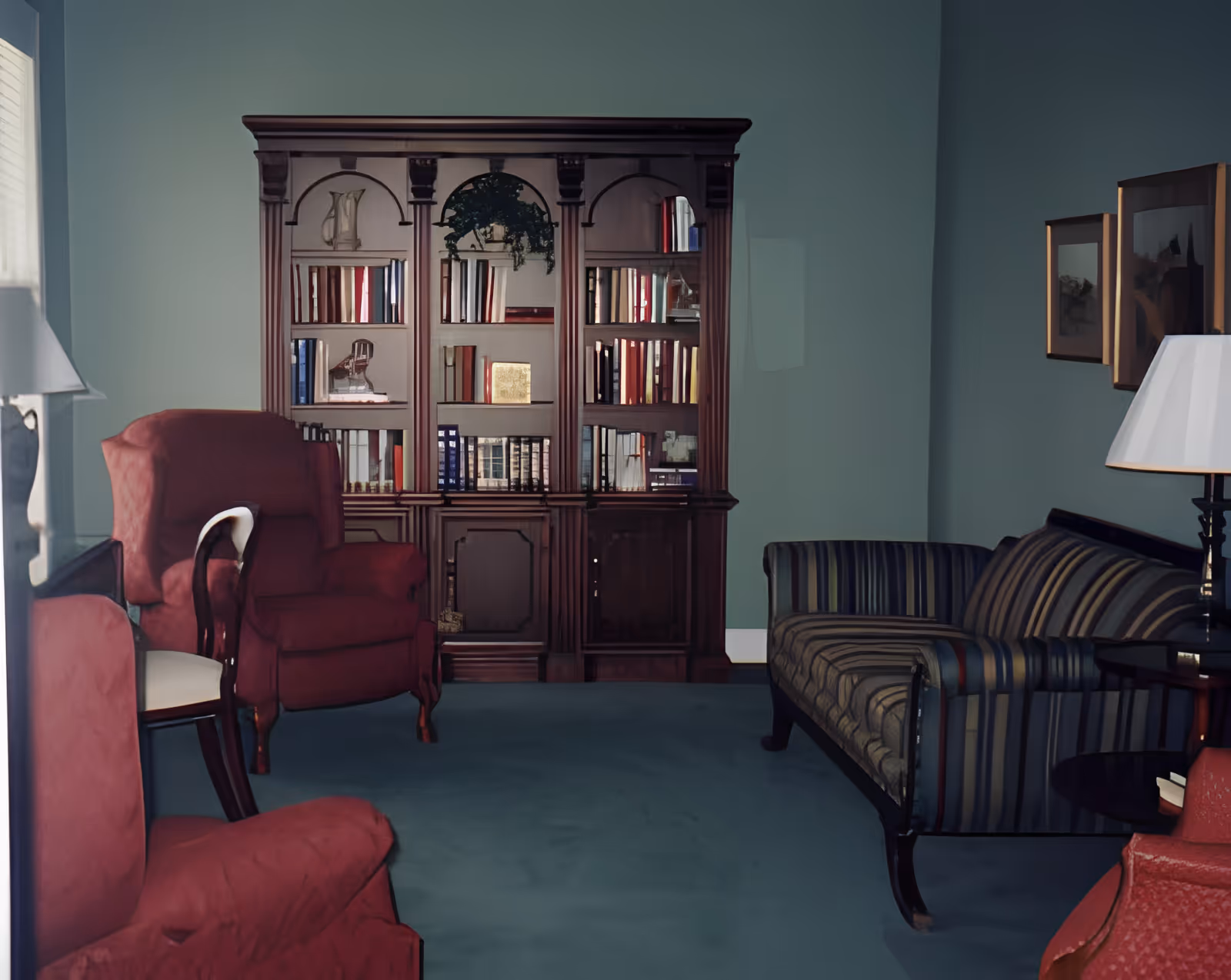 A cozy living room with a dark wooden bookshelf filled with books and decorative items against a teal wall. The room features two red upholstered armchairs, a striped sofa, a floor lamp with a white shade, and framed artwork on the wall.