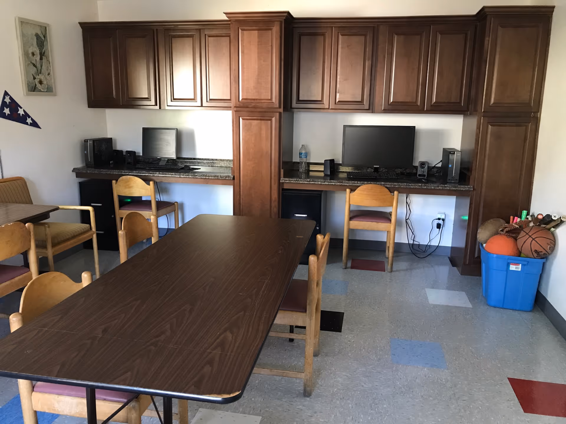A room with a long wooden table surrounded by wooden chairs with purple cushions. Along the back wall, there are two computer workstations with monitors, keyboards, and desktop towers placed on a countertop beneath wooden cabinets. To the right, a blue bin contains sports equipment including basketballs and other balls. The floor has a pattern of square tiles in various colors including gray, blue, red, and black. A framed picture and a triangular wall decoration are visible on the left wall.
