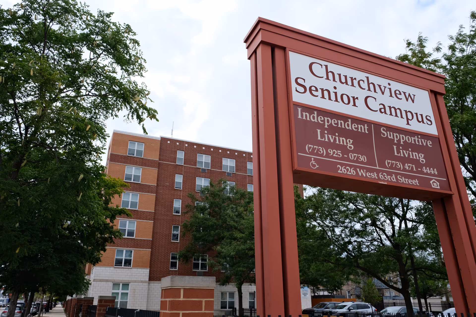 Outdoor view of Churchview Senior Campus sign with contact information for Independent Living and Supportive Living, with a multi-story brick building and trees in the background.