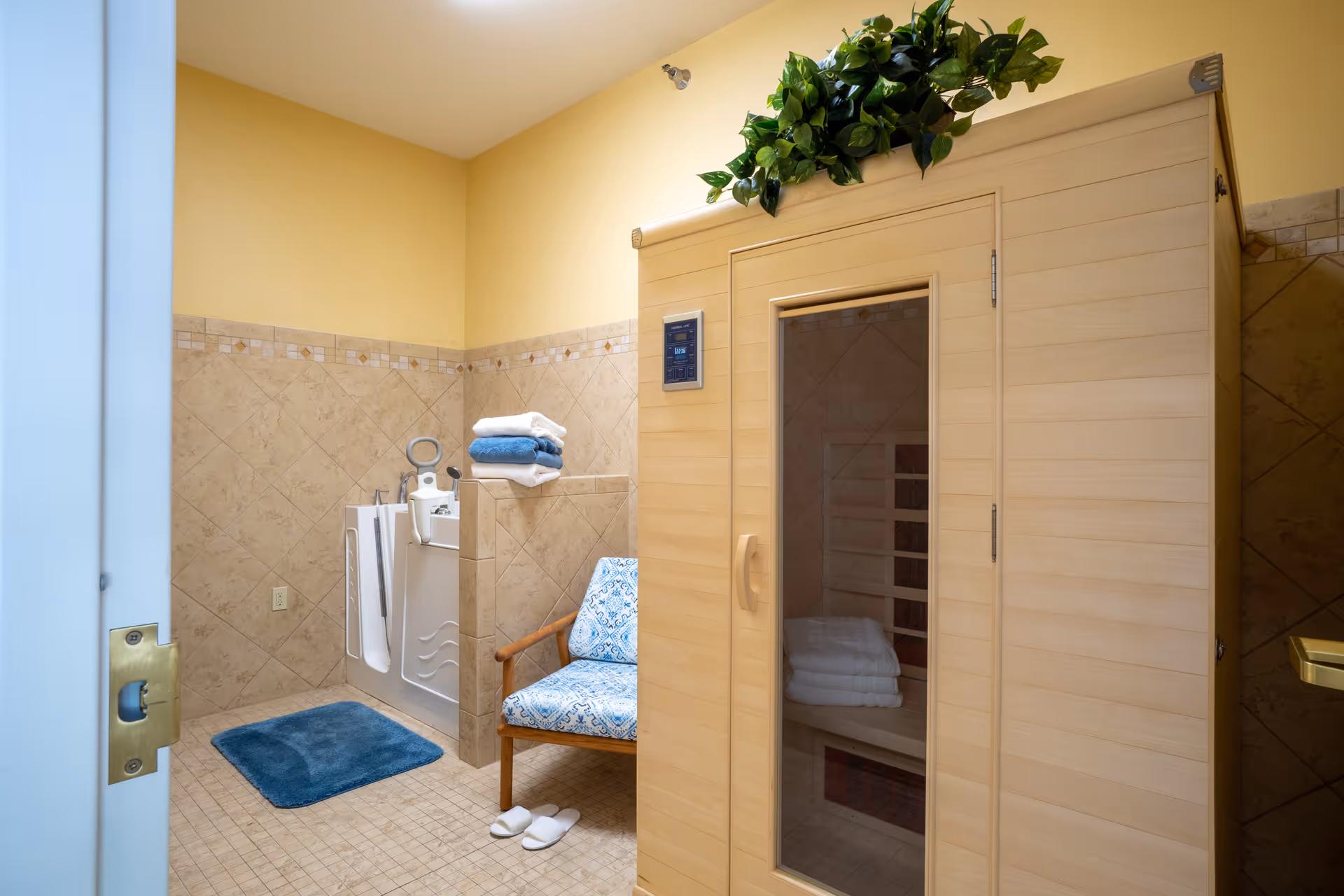 A room with a wooden sauna on the right side, a blue patterned chair with wooden arms, a stack of towels on a tiled ledge, a blue bath mat on the floor, and a walk-in bathtub with safety handles. The walls are tiled halfway up with beige tiles and painted yellow above.