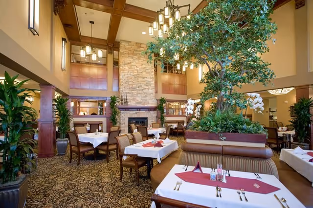 A spacious dining room with tables covered in white tablecloths and red napkins, surrounded by wooden chairs. The room features a large stone fireplace, tall ceilings with wooden beams, hanging light fixtures, and a large indoor plant centerpiece. The carpet has an intricate pattern, and there are several potted plants along the walls.