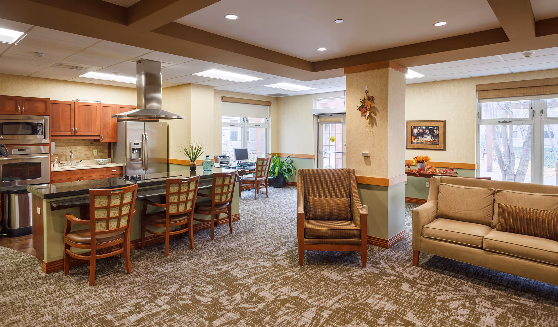 A spacious and well-lit common area in a senior living facility featuring a kitchen with wooden cabinets, stainless steel appliances, and a black countertop island with four chairs. Adjacent to the kitchen is a seating area with a brown armchair and a beige sofa. Large windows allow natural light to fill the room, and there are plants and framed artwork on the walls.