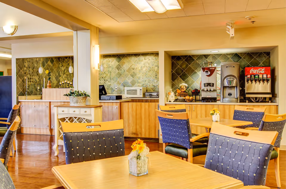 A bright dining area with wooden tables and chairs featuring blue cushioned seats. The background shows a beverage station with a coffee machine, soda dispenser, microwave, and toaster oven against a tiled backsplash. Small flower arrangements are placed on the tables.
