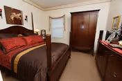 A cozy bedroom with a wooden four-poster bed dressed in red and brown bedding. There is a wooden armoire against the wall, a dresser with a mirror, and a small window with a white curtain. The walls are light-colored with a decorative border near the ceiling, and a framed painting hangs above the bed.