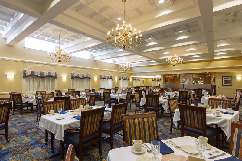 A spacious dining room with multiple tables covered in white tablecloths, each set with cups, plates, glasses, and silverware. The room features striped upholstered chairs, patterned carpet, chandeliers hanging from a coffered ceiling, and windows with valances along the walls.