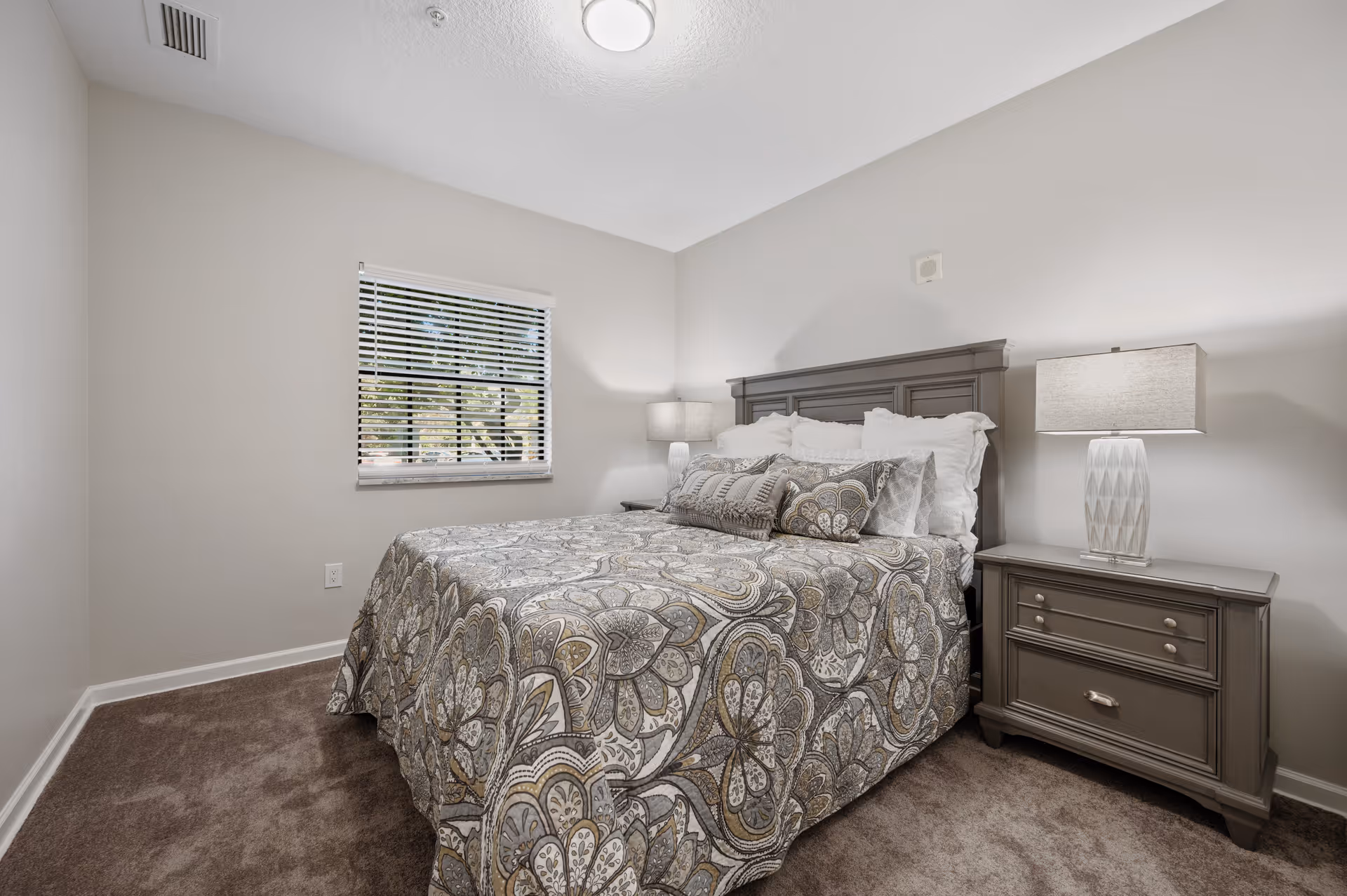 A bedroom with a neatly made bed featuring a patterned bedspread and multiple pillows. There is a window with blinds on the left wall, and two matching bedside tables with lamps on either side of the bed. The walls are painted light gray and the floor is carpeted in brown.