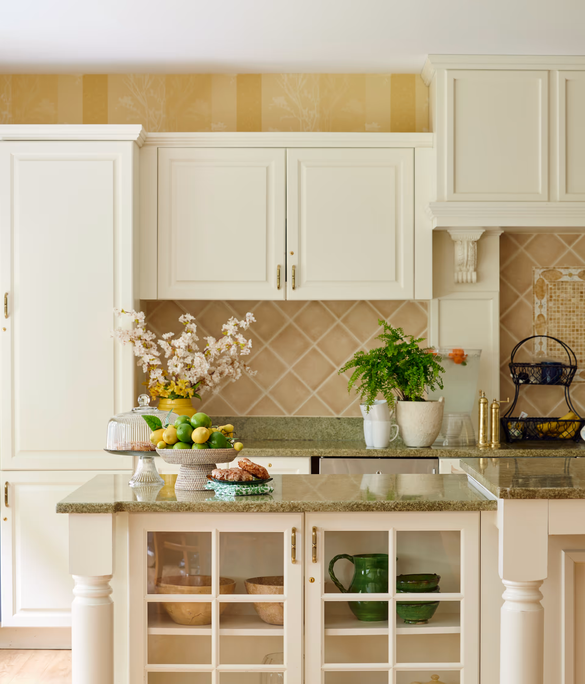 A bright kitchen with cream-colored cabinets and a green granite countertop. The kitchen island features glass-front cabinets displaying bowls and a green pitcher. On the countertop, there is a bowl of green apples and lemons, a plate with cookies, a glass cake stand with a cover, a vase with flowers, and a potted fern. The backsplash is tiled in a beige diamond pattern.