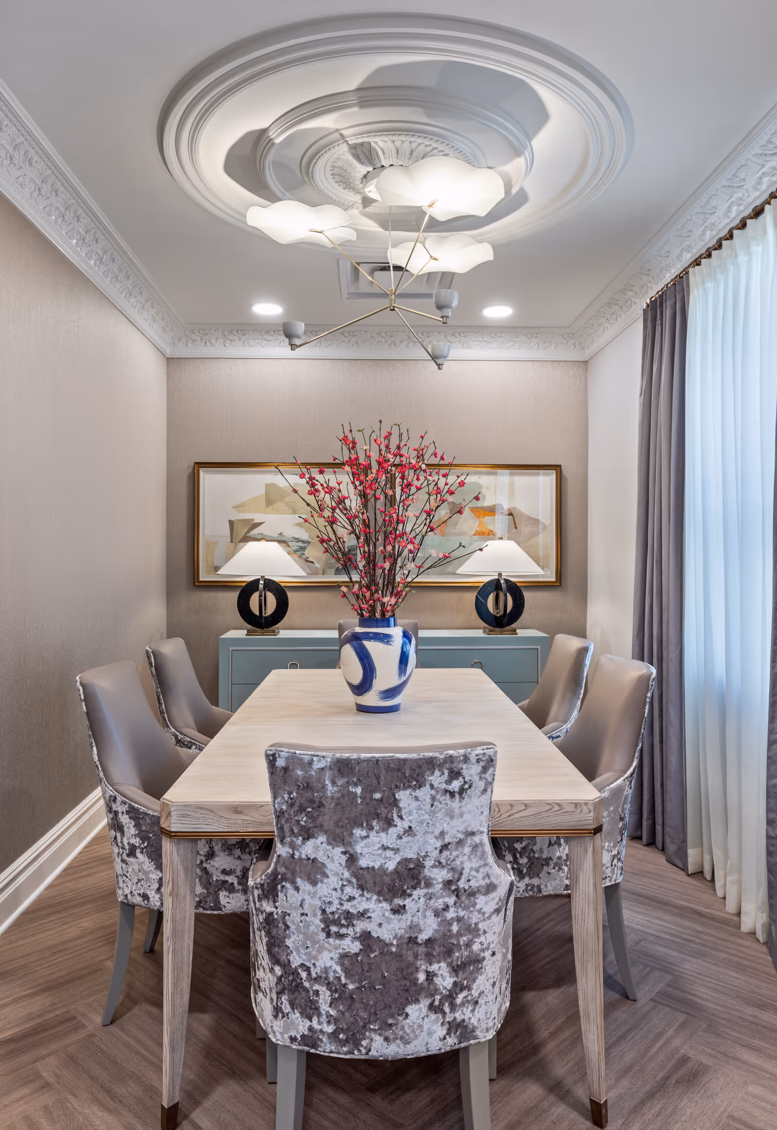 A stylish dining room with a rectangular wooden table surrounded by six upholstered chairs with a textured gray and white pattern. On the table is a blue and white vase holding branches with pink blossoms. Behind the table is a light blue sideboard with two modern black and white lamps and a large abstract painting on the wall. The room features ornate crown molding, a decorative ceiling medallion with a modern chandelier, and floor-to-ceiling curtains on the right side.