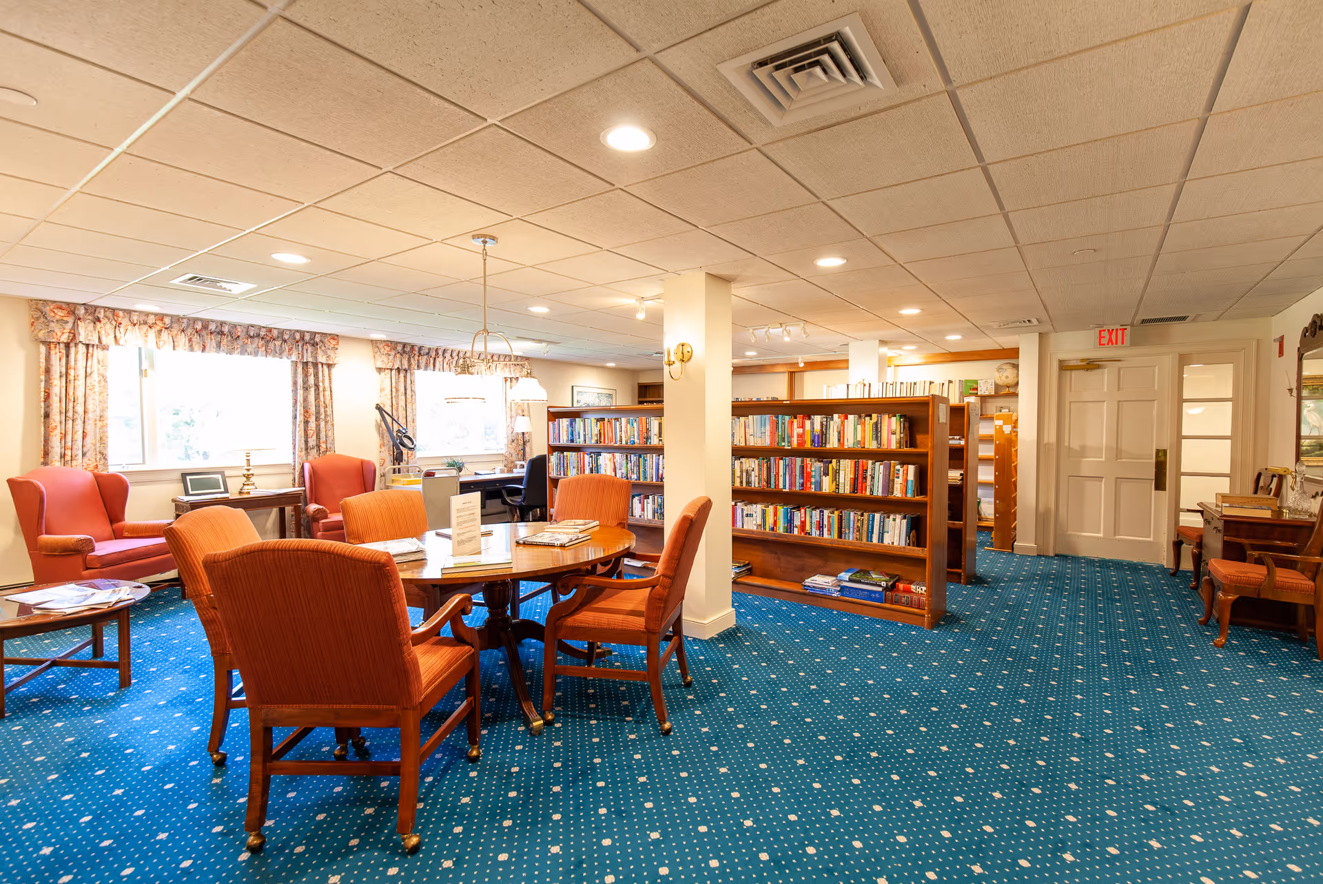 A cozy library or reading room in a senior living facility with blue carpet, wooden bookshelves filled with books, a round wooden table surrounded by four orange upholstered chairs, and two orange armchairs near windows with floral curtains. The room is well-lit with ceiling lights and has a calm, inviting atmosphere.