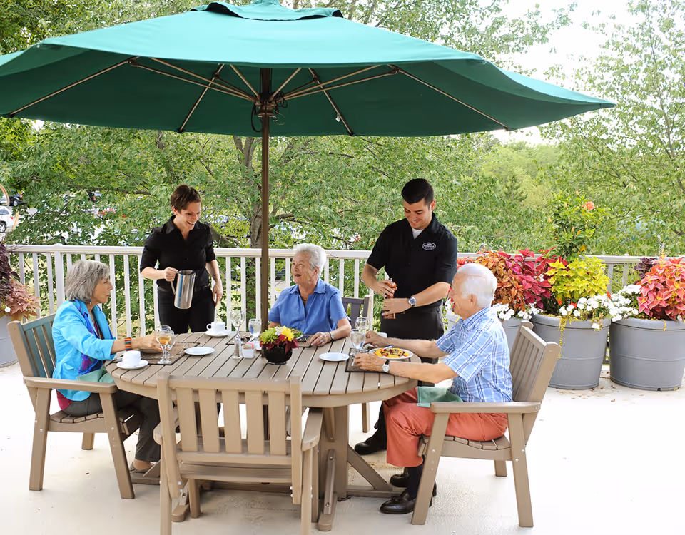 Three elderly people sitting around a round outdoor table under a large green umbrella, with two staff members serving them drinks. The setting is a patio with potted plants and trees in the background.