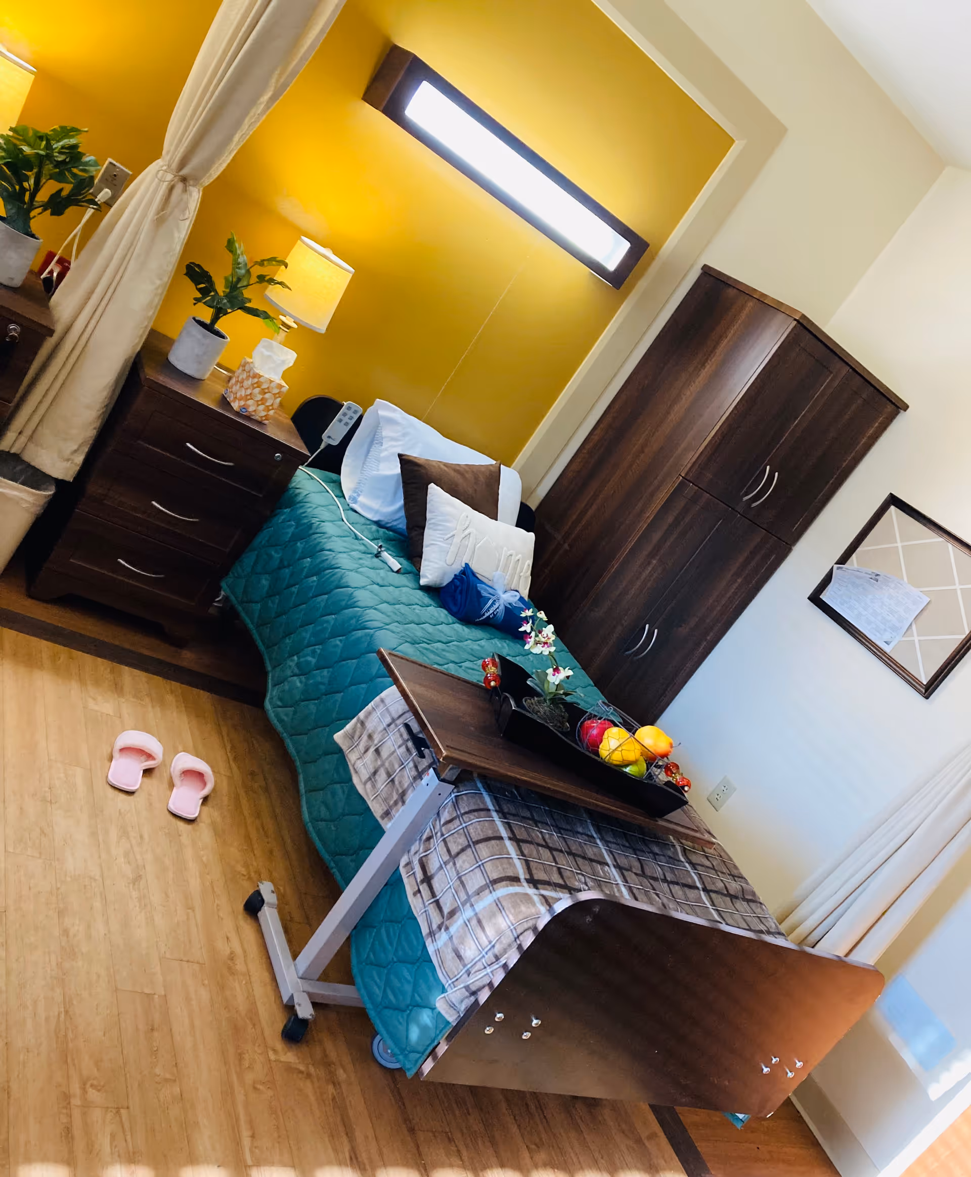 A cozy bedroom in a senior living facility with a single bed covered in a teal quilt and plaid blanket. The bed has pillows and a tray table with a fruit bowl and flowers on it. There is a wooden nightstand with a lamp, tissue box, and potted plant next to the bed. A wooden wardrobe and a framed bulletin board are on the wall. Pink slippers are on the wooden floor near the bed.