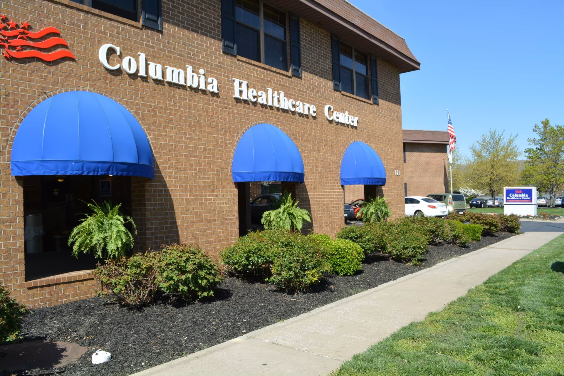 Exterior view of Columbia Healthcare Center, a brick building with three blue awnings over windows and a door. There are green bushes and plants along the building, a sidewalk, and a parking lot with cars and an American flag in the background.