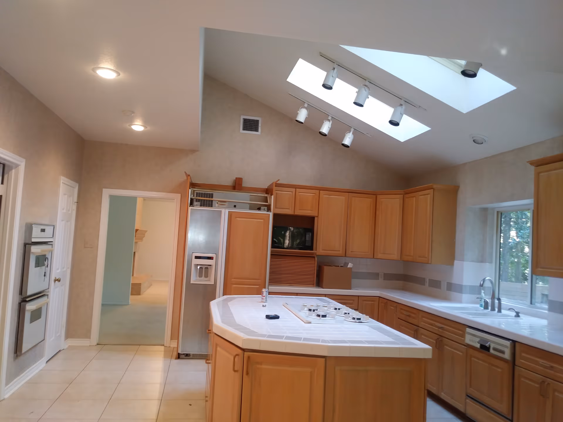 A spacious kitchen with wooden cabinets, a large island with a built-in stovetop, a double oven, a refrigerator with a water dispenser, a dishwasher, and a window above the sink. The ceiling features two skylights and track lighting.