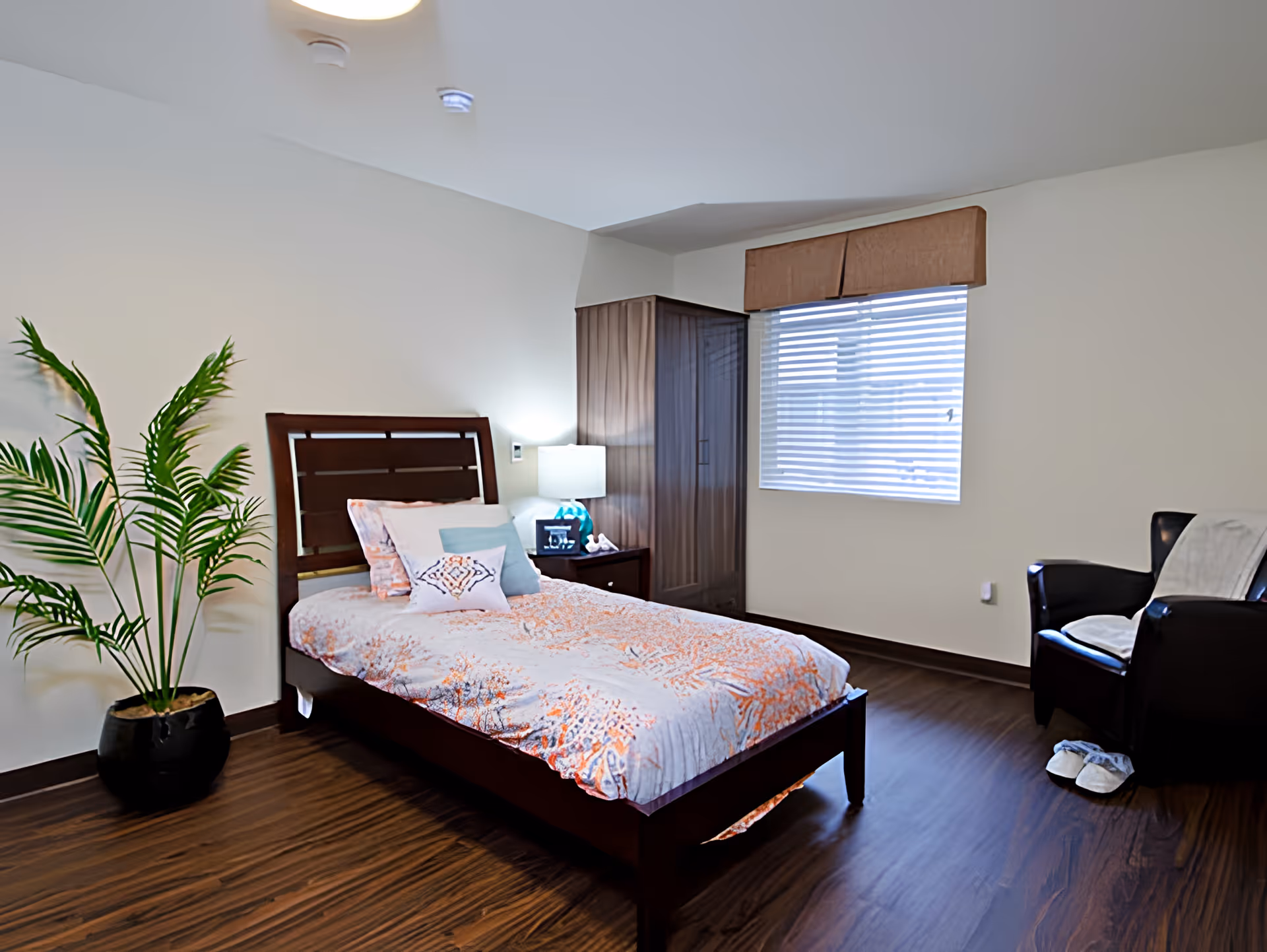 A neatly arranged bedroom with a single bed featuring a floral patterned bedspread and multiple pillows. Next to the bed is a nightstand with a lamp and a small decorative item. A large potted plant is placed on the left side of the bed. There is a window with blinds and a brown valance on the right wall. A dark wooden wardrobe stands in the corner, and a black armchair with a white throw blanket and slippers on the floor is positioned near the window. The room has wooden flooring and light-colored walls.