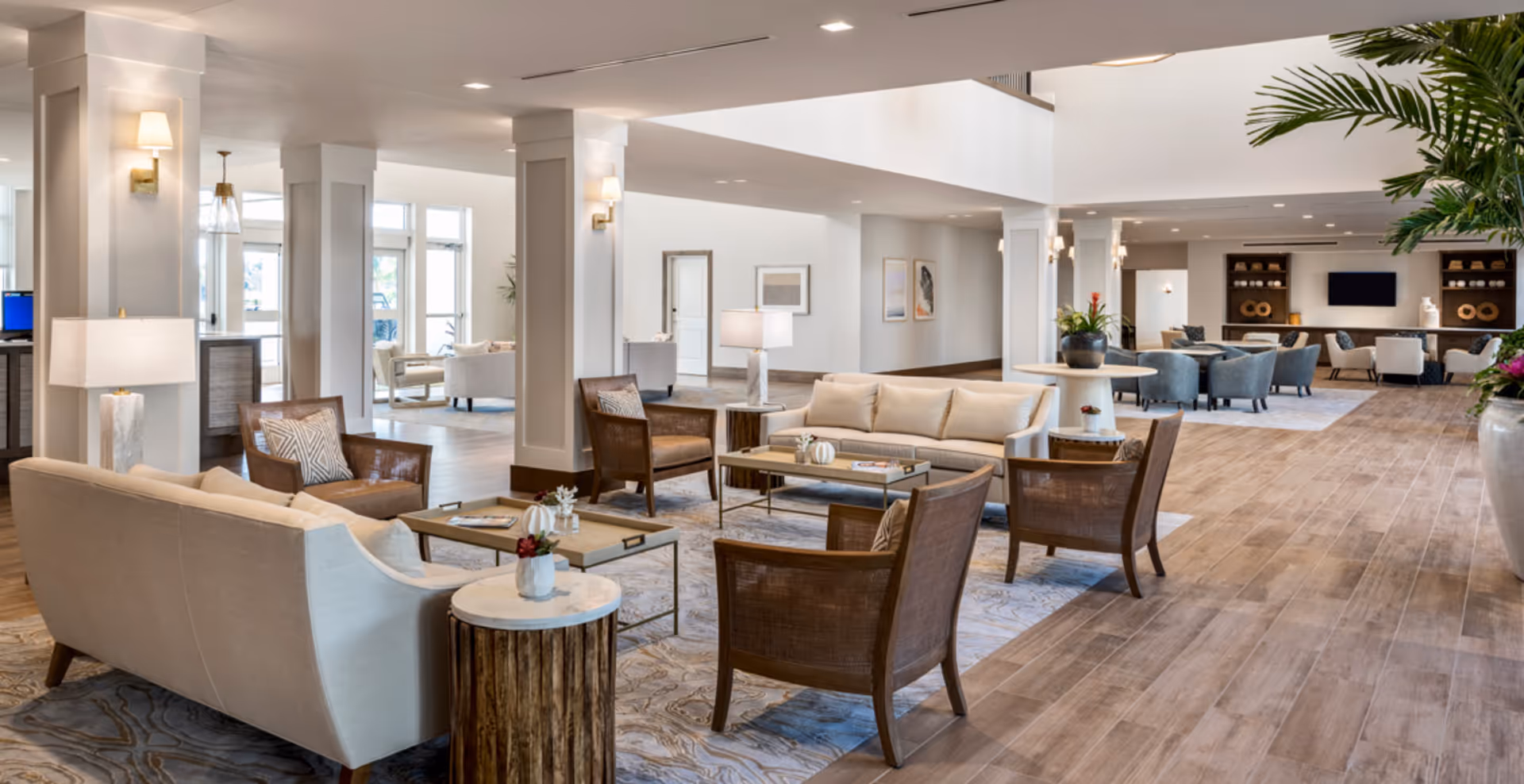 A spacious and well-lit senior living facility common area with multiple seating arrangements including beige sofas, wooden armchairs with cushions, coffee tables, and side tables with lamps and small plants. The room features wooden flooring, white walls, large windows, and decorative wall art. There are also plants and a TV mounted on the far wall.