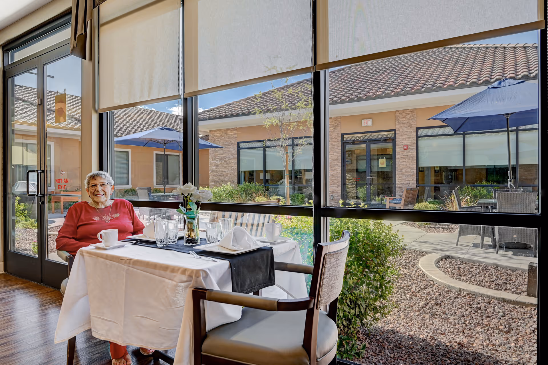 An elderly woman in a red sweater sits at a set dining table by large windows overlooking a sunny courtyard with patio umbrellas.