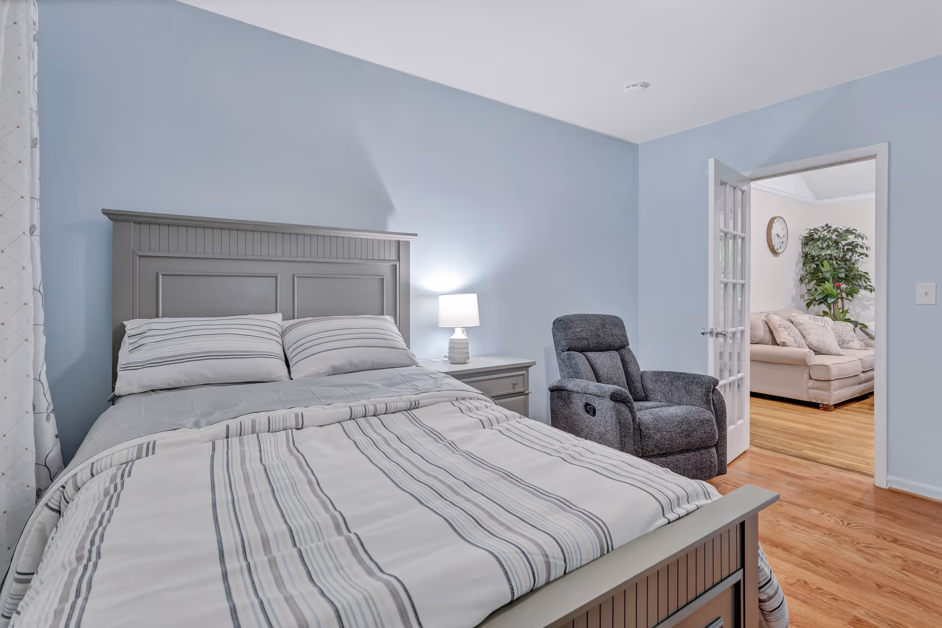A bedroom with a gray wooden bed frame and striped bedding. Next to the bed is a nightstand with a white lamp. A gray recliner chair is positioned near an open door that leads to a living room with a beige sofa, a clock on the wall, and a large green plant. The walls in the bedroom are painted light blue and the floor is wooden.