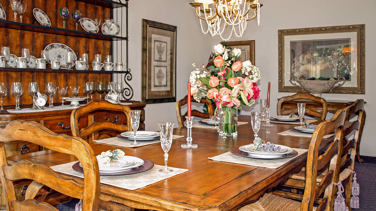 A wooden dining table set for six with plates, crystal glasses, and floral napkin holders. A vase with a colorful bouquet of flowers and two red candles in glass holders are in the center of the table. Behind the table is a wooden hutch displaying fine china and glassware. The room has framed artwork on the walls and a chandelier hanging above the table.