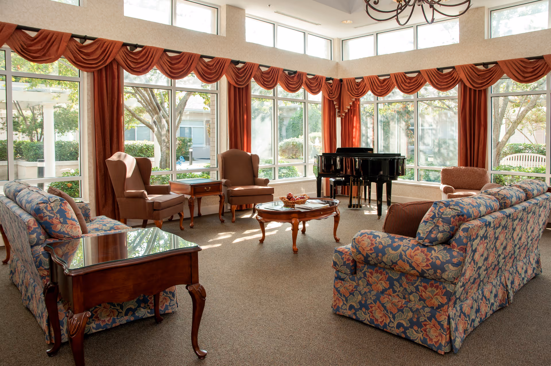 Sunlit communal lounge with floral sofas, armchairs, a grand piano and large windows overlooking a garden.