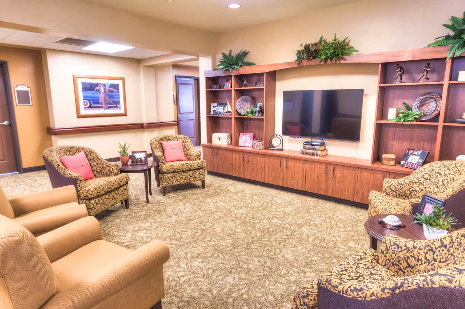 A cozy living room area in Catalina Springs Memory Care featuring patterned armchairs with pink cushions, beige armchairs, a large wooden entertainment center with a flat-screen TV, decorative plants, and various ornaments and framed pictures on the shelves. The room has beige walls, carpeted flooring with a floral pattern, and a framed picture on the wall near a door.