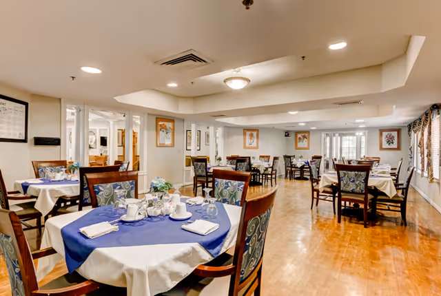Dining room with round and rectangular tables set with tablecloths and place settings in a senior living facility.