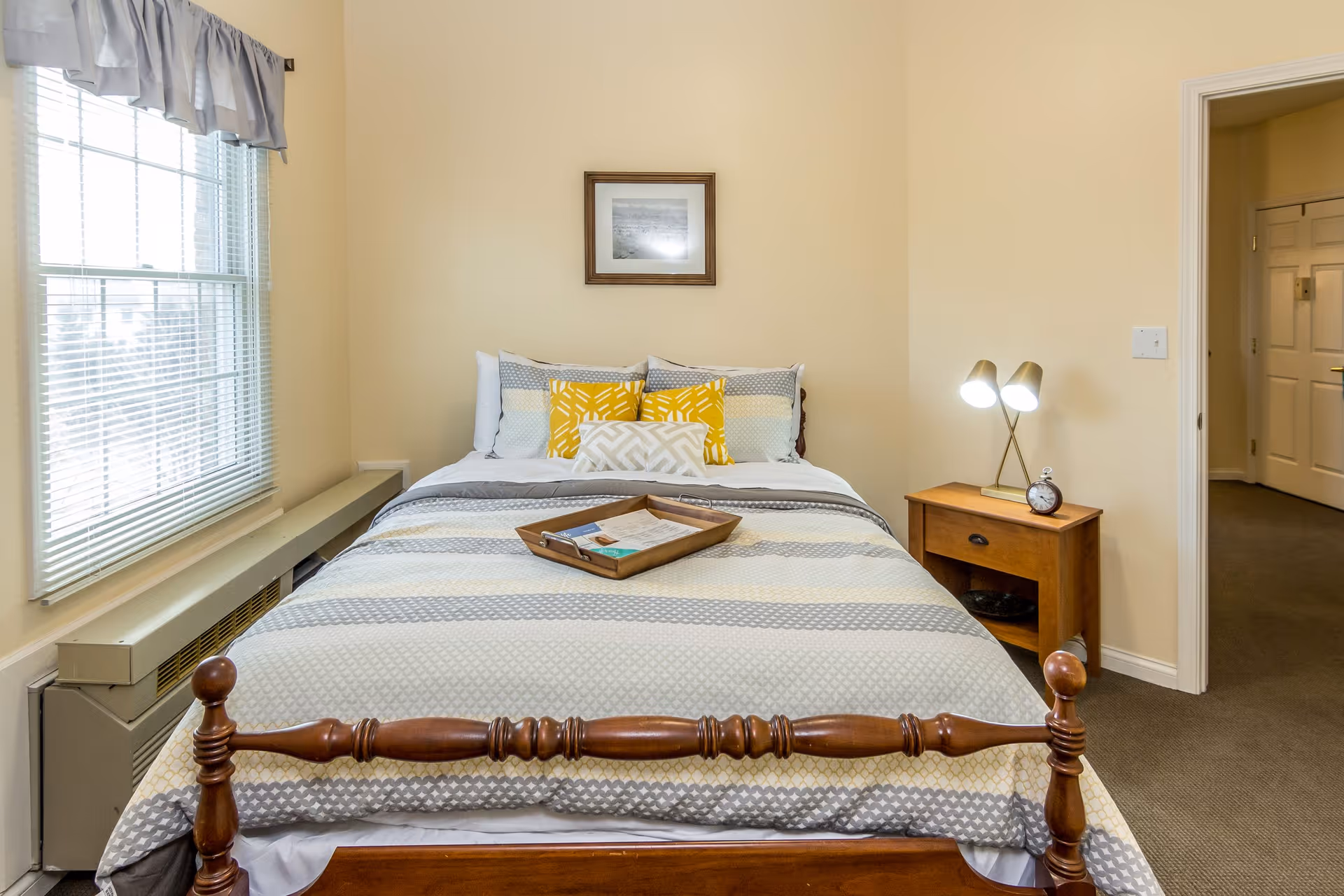 Neatly made bed in a softly lit bedroom with a window on the left, a wooden bedside table and lamp on the right.