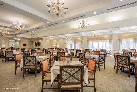 A spacious dining room with multiple tables covered with white tablecloths, each surrounded by wooden chairs with patterned upholstery. The room is well-lit with large windows allowing natural light and elegant chandeliers hanging from the ceiling. The decor is warm and inviting, suitable for a senior living facility.