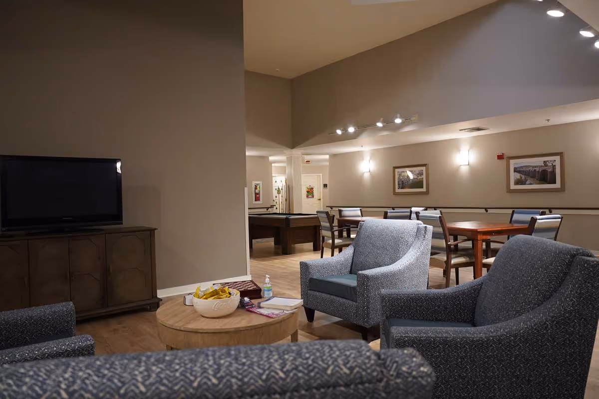 A cozy common area in a senior living facility featuring three upholstered armchairs around a round wooden coffee table with a bowl of snacks, books, and hand sanitizer. In the background, there is a wooden cabinet with a flat-screen TV on top, a pool table, and a dining area with tables and chairs. The walls are decorated with framed pictures and soft lighting fixtures.